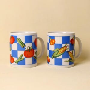 May include: Two white ceramic mugs with a blue and white checkered pattern. Each mug is decorated with illustrations of vegetables, including tomatoes, asparagus, corn, and onions. The mugs have white handles.
