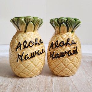 以下が含まれることがあります： パイナップルの形をした塩コショウ入れのペア。塩コショウ入れは黄色で、表面はざらざらしており、上部は緑色です。「Aloha Hawaii」の文字が黒い筆記体で各入れ物の前面に書かれています。