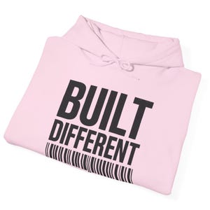 Peut inclure: Un sweat-shirt à capuche rose clair avec le texte noir BUILT DIFFERENT imprimé dans une police sans empattement grasse, placé au-dessus d'un graphique de code-barres noir. Le vêtement comprend une capuche assortie avec des cordons de serrage.
