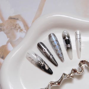 Op de afbeelding: Vijf lange, amandelvormige kunstnagels met ingewikkelde ontwerpen. De nagels hebben een verloop van zwart naar wit, zilveren accenten en oogachtige details. Ze zijn gerangschikt op een witte, ovaalvormige schaal.