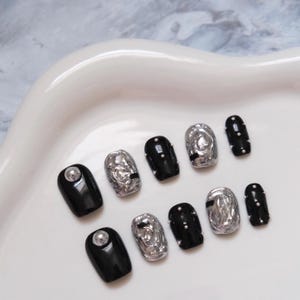 Puede incluir: Un conjunto de diez uñas artificiales en una variedad de diseños. Algunas uñas son negras sólidas con un tachuela plateada, mientras que otras son plateadas con una apariencia texturizada. Varias uñas son negras con pequeños acentos blancos perlados.