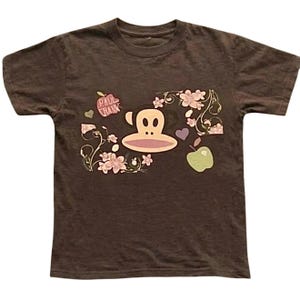 Könnte beinhalten: Ein braunes Kurzarm-T-Shirt mit einer Grafik eines Affengesichts, rosa Blumen, einem lila Herz und zwei Äpfeln. Ein Apfel ist rot mit dem aufgedruckten Text PAUL FRANK, der andere ist hellgrün. Das Motiv ist mittig auf der Brust platziert.