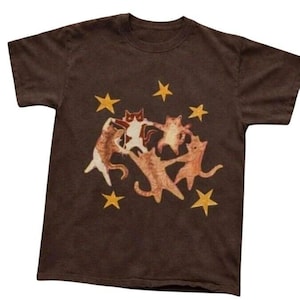 May include: A dark brown short-sleeve t-shirt featuring a graphic print of four cats holding hands in a circle. The cats are depicted in shades of orange, brown, and white. Five yellow stars are scattered around the cats on the front of the shirt.