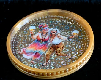 May include: A round, amber-colored resin tray featuring a central graphic of two individuals wearing sunglasses and headwear, each holding a wine glass. The tray surface is embedded with shimmering, iridescent, leopard-print-like patterns.
