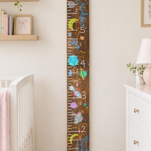 May include: A tall, dark brown wooden growth chart featuring painted space-themed illustrations including planets, stars, rockets, and an astronaut. The board displays white measurement markings and numbers from one to six, with the name Jackson and the text loved beyond measure written in blue script.