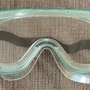 May include: Clear safety goggles with a light green frame and a black elastic strap. The goggles have a wide, curved design to protect the eyes. The lenses are transparent, allowing for clear visibility.