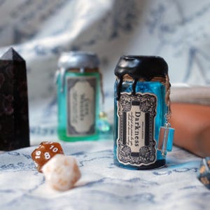 May include: A blue potion bottle with a black wax seal and a label that reads "Darkness". The bottle has a decorative charm attached. Other bottles and dice are in the background. The potion bottle is approximately 4 inches tall.