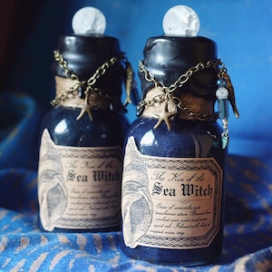 May include: Two dark blue glass bottles with a sparkling liquid, labeled "The Kiss of the Sea Witch." Each bottle has a black cap, decorative chain, starfish charm, and a clear, spherical stopper. The labels feature a seashell illustration.