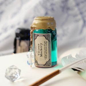 Può includere: Una piccola bottiglia di vetro riempita di liquido verde acqua, sigillata con un tappo di cera dorata e uno spago. L'etichetta recita "Vicious Mockery" con testo aggiuntivo. Un ciondolo di cristallo trasparente pende dalla bottiglia.