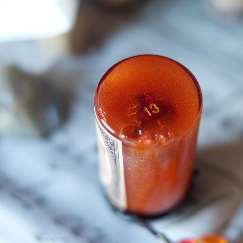 May include: A cylindrical, amber-colored candle with a gold number 13 embedded in the wax. The candle has a glossy finish and is partially filled with a reddish-brown liquid. The label is partially visible, with a decorative border.