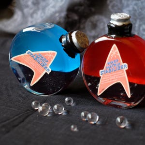 May include: Two round glass bottles filled with blue and red liquid, each featuring a star-shaped label that reads Warp Matrix Stabilizer. The bottles have silver-colored caps and are surrounded by several small, clear, spherical glass beads.