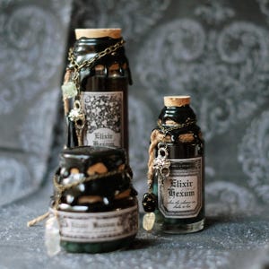 Könnte beinhalten: Drei Glasflaschen mit Korken und schwarzen Wachssiegeln. Jede Flasche hat ein Etikett mit der Aufschrift "Elixir Hexum". Die Flaschen sind mit Ketten, Charms und Kristallen verziert. Die Gesamtästhetik ist dunkel und mystisch.