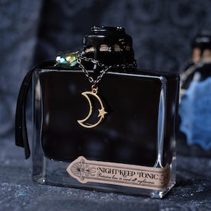 May include: A black square glass bottle with a black stopper and a gold crescent moon and star pendant. The bottle has a label that reads "NIGHT KEEP TONIC Protective brew to ward off nightmares."