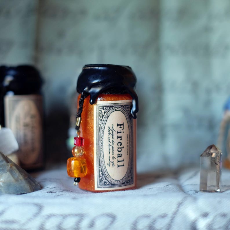 May include: A small, orange-filled glass bottle with a black wax seal and a decorative label that reads "Fireball." The label also includes the text "conjured dice potion to get, check and decision making." A beaded charm hangs from the bottle.