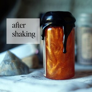 May include: A small glass bottle filled with a shimmering, golden-orange liquid. The bottle is sealed with a black wax top that has dripped down the sides. The text "after shaking" is in a white box in the upper left corner. The bottle is on a white surface.