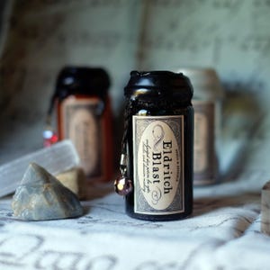 May include: A dark glass bottle with a black wax seal and a decorative label that reads "Eldritch Blast". The label also includes the text "Spell & Spindle" and "enchanted dice potion for spells, hexes and illusion making". A small charm hangs from the bottle.