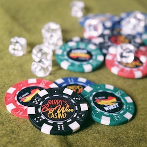 May include: A collection of casino chips in red, green, blue, and black, with the text "Barry's Big Win Casino" on one chip. Clear dice are scattered around the chips on a green surface. The chips have various designs.