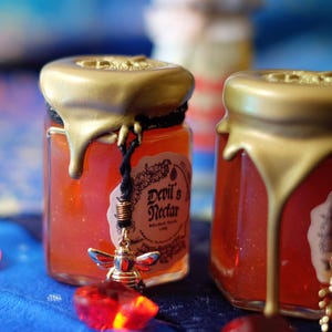 May include: Two small glass jars filled with a red liquid and topped with gold lids. The jar on the left has a label that reads "Devil's Nectar" and a small bee charm hanging from the lid.
