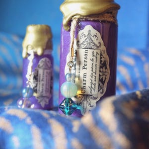 May include: Two small, purple potion bottles with gold wax seals and decorative tags. The tags read "Charm Person" and "enchanted dice potion for rolls, checks, and decision making." Each bottle has a beaded charm with blue and clear beads.