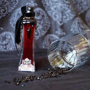 May include: A tall, rectangular glass bottle filled with a deep red liquid, topped with black wax and a black velvet ribbon. The bottle features a small black coffin-shaped emblem and a label reading Blue Blood. A clear glass tumbler lies nearby.