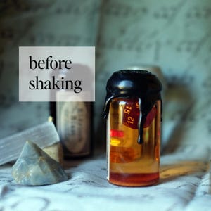 May include: A small glass bottle filled with a golden liquid and a red die. The bottle is sealed with black wax, with the text "before shaking" in a white box. The background includes other bottles and crystals.