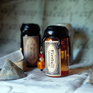 May include: A small, clear glass bottle filled with an orange liquid, sealed with black wax and a decorative label that reads "Fireball". A charm with red and orange beads hangs from the bottle. Other bottles are visible in the background.