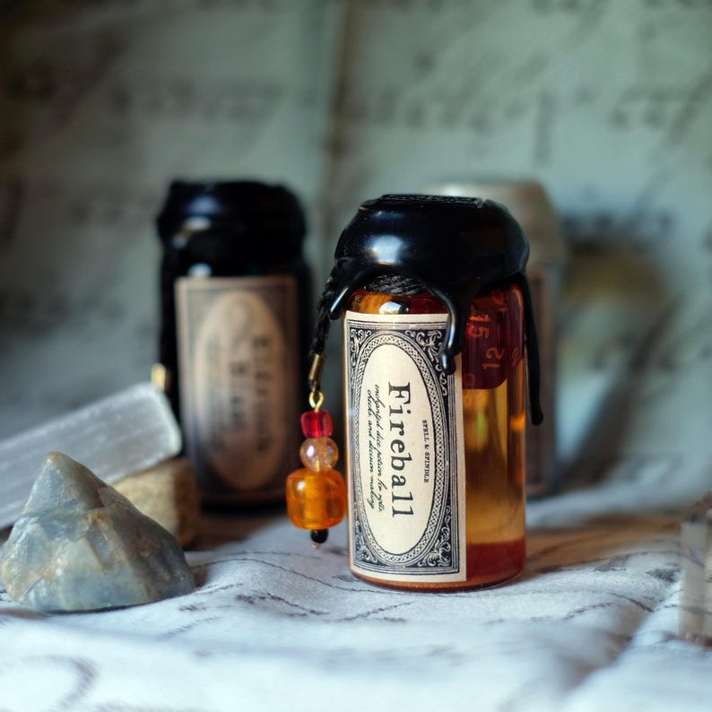 May include: A small, clear glass bottle filled with an orange liquid, sealed with black wax and a decorative label that reads "Fireball". A charm with red and orange beads hangs from the bottle. Other bottles are visible in the background.