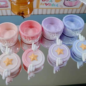 May include: A collection of pastel-colored ceramic containers and lids. The containers are round with decorative edges, and the lids feature a star design with angel wings. Colors include pink, lavender, and blue.