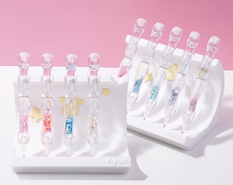 May include: Set of six clear glass pens with colorful glitter and gold stars inside. The pens are displayed in two white holders with the word "Ayun" printed on them.