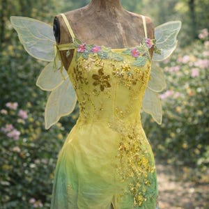 以下が含まれることがあります： 黄色と緑色の妖精のドレス。花柄とスパンコールの装飾が施されています。フィットした胴体、流れるようなスカート、透明な翼が付いています。庭に置かれたヴィンテージのマネキンにドレスが飾られています。