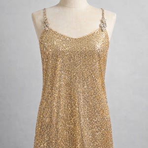 May include: A gold sequin camisole top with a V-neck and delicate jeweled straps. The shimmering top is displayed on a mannequin, showcasing its intricate design and overall elegance. This glamorous piece is perfect for special occasions.