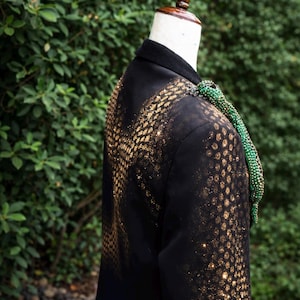 May include: Black blazer with gold glitter pattern on the back and sleeves. A green beaded embellishment drapes over the shoulder. The jacket is displayed on a mannequin against a backdrop of green foliage.