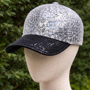 May include: A silver sequin baseball cap with a black brim. The cap is displayed on a mannequin head against a backdrop of green foliage. The cap's crown is covered in shiny silver sequins.