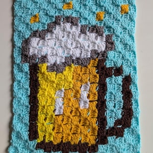 May include: Crocheted square featuring a pixelated beer mug design. The mug is yellow with brown outlines and a white foam top. The background is light blue with yellow accents. Handmade craft item.