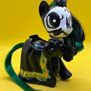 May include: A black and green painted pony figurine with a skull and crossbones design. The pony has a green mane and tail and is wearing a black dress with gold accents.