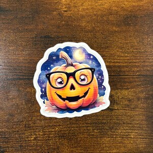 May include: A colorful sticker featuring a smiling pumpkin wearing black glasses. The pumpkin is orange with a carved jack-o'-lantern face. The background shows a night sky with stars and a crescent moon, with fall leaves at the bottom.