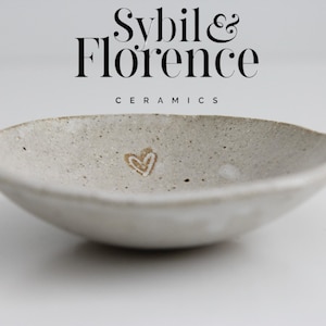 May include: A small, off-white ceramic bowl with a speckled texture and a heart design. The bowl has a shallow, rounded shape and is branded with the text "Sybil & Florence Ceramics".