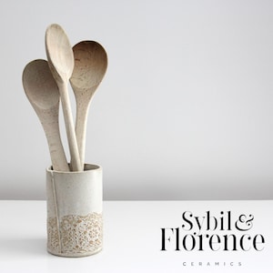 May include: A ceramic utensil holder containing three wooden spoons. The holder is off-white with a textured, lace-like pattern at the base. The spoons are light-coloured wood. The text "Sybil & Florence Ceramics" is in the lower right corner.
