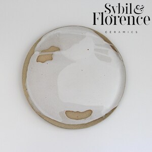 May include: A circular ceramic plate featuring a natural tan clay base partially covered with a glossy white glaze. The glaze application is uneven, revealing patches of the raw, speckled tan surface. The text Sybil & Florence CERAMICS is visible.