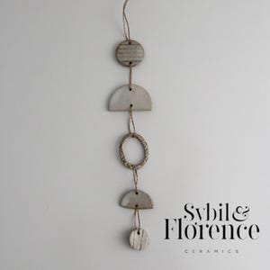 May include: A ceramic wall hanging with a series of textured, off-white shapes: circles, a half-circle, and a ring, connected by twine. The design includes the text "Sybil & Florence Ceramics" in a stylish font.