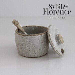 May include: A speckled light grey ceramic jar with a matching lid and a small spoon. The jar features a textured finish and a natural clay rim. The text Sybil & Florence CERAMICS is printed in black in the upper right corner of the image.