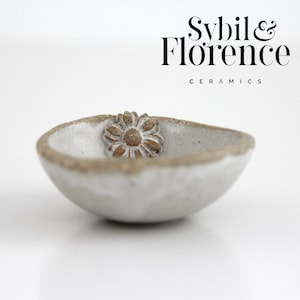 May include: A small, off-white ceramic bowl with a textured, uneven rim and a brown, embossed daisy flower detail on the interior wall. The text Sybil & Florence CERAMICS is printed in black in the upper right corner of the image.