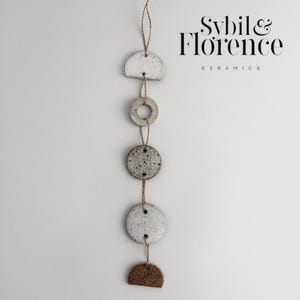 May include: A ceramic wall hanging featuring a series of white and speckled grey shapes connected by twine. The shapes include a half-circle, a ring, two circles, and a half-circle with a textured design. The text "Sybil & Florence Ceramics" is visible.
