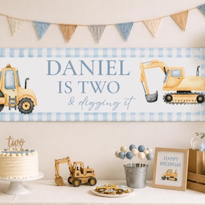 May include: A rectangular birthday banner featuring a light blue gingham border, a yellow steamroller, a yellow excavator, and the text DANIEL IS TWO & digging it in blue lettering. The banner is displayed above a table with party decorations.