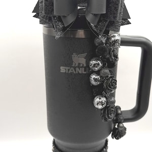 May include: Black Stanley tumbler with a handle, adorned with a large bow, glitter accents, and a decorative charm of roses and beads. The tumbler has a black base with a decorative band at the bottom.