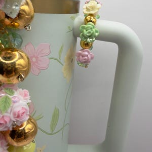 May include: A light blue floral-patterned tumbler with a gold rim and a white handle. The tumbler is decorated with pink and yellow flowers and gold ornaments. A decorative bracelet with pink, yellow, and green flower beads hangs from the handle.