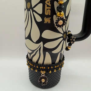 May include: A black and cream-colored Stanley tumbler with a floral design, adorned with beaded embellishments in black, gold, and pearl. A decorative flower charm hangs from the base. The handle is black.