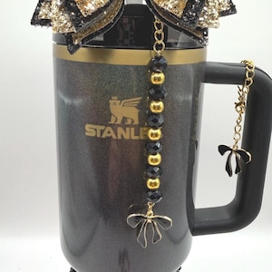 May include: A black and gold Stanley tumbler with a black handle, adorned with gold and black bows, and decorative charms. The tumbler features the Stanley logo and is embellished with gold and black beads.