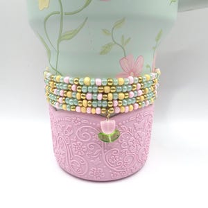 May include: A light green floral tumbler with a pink base and a beaded wrap. The wrap features yellow, pink, and green beads, with a dangling tulip charm. The tumbler has a floral design.