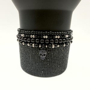 May include: A stack of four beaded bracelets featuring glossy black round beads, metallic silver-toned spacers, and small clear crystal accents. A single dark, textured skull-shaped charm hangs from the bottom bracelet in the stack.